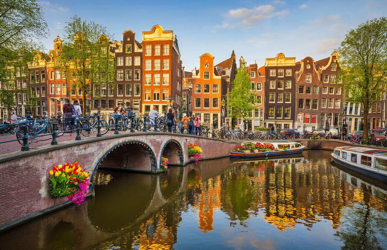 Create a realistic image of a vibrant Amsterdam canal scene featuring colorful Dutch row houses reflected in calm water, with a traditional arched bridge in the foreground adorned with bicycles, warm golden afternoon sunlight casting a welcoming glow over the scene, a few Indian tourists enjoying the view and taking in the surroundings, tulips and flower boxes adding pops of color, the atmosphere feeling lively yet cozy and familiar, with houseboats gently floating along the canal, capturing the diverse and warm character of the city. Absolutely NO text should be in the scene.
