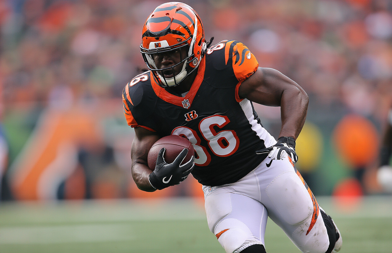 Create a realistic image of a black male NFL running back in action during a game, wearing a Cincinnati Bengals uniform, displaying powerful running form with muscular build, carrying a football while breaking through the defensive line, showing his physical strength and determination, set on a professional football field with stadium lighting and crowd in the background, capturing the intensity and athleticism of professional football, absolutely NO text should be in the scene.