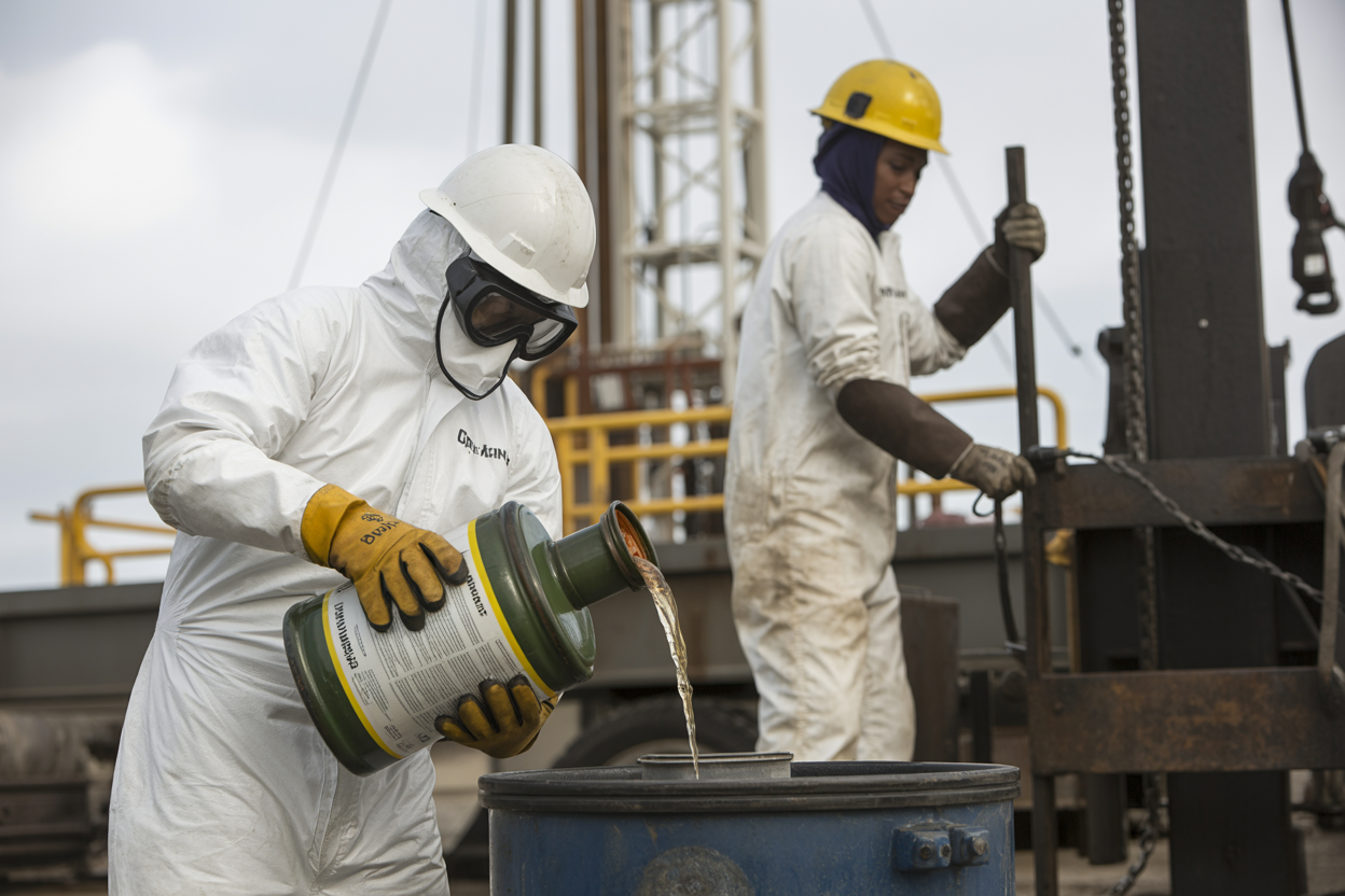 Create a realistic image of oil drilling rig workers in protective gear handling chemical containers and materials, showing a white male worker in full hazmat suit and safety goggles carefully pouring chemicals from a labeled container while a black female worker in hard hat and protective clothing operates material handling equipment in the background, industrial drilling rig environment with metal structures and equipment visible, serious safety-focused atmosphere with proper lighting to emphasize the hazardous nature of chemical handling operations, absolutely NO text should be in the scene.