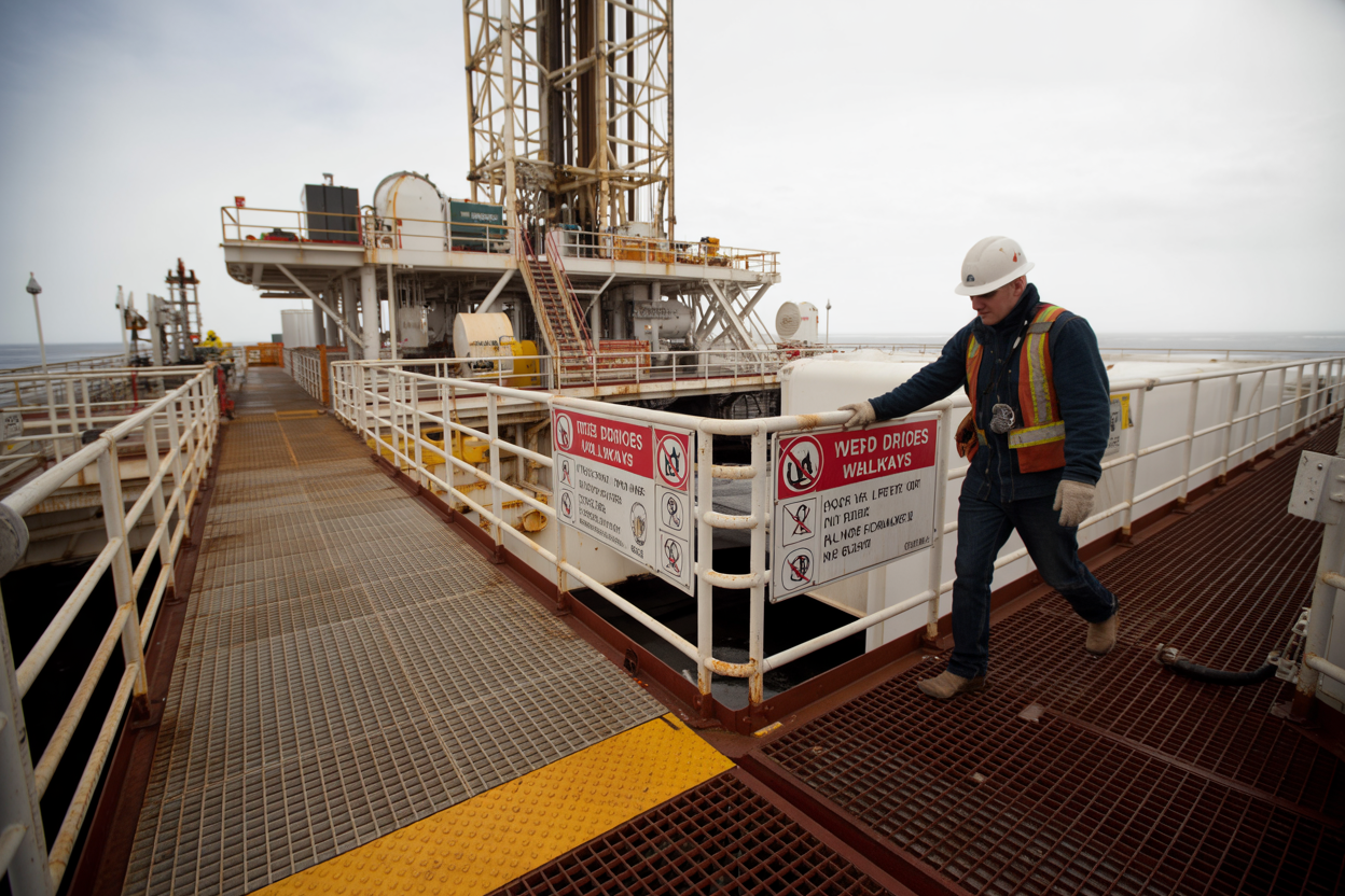 Create a realistic image of a drilling rig platform with safety warning signs about wet surfaces and proper walkways, showing industrial metal grating flooring with some areas having yellow anti-slip tape, safety railings along elevated walkways, a white male worker in hard hat and safety vest carefully walking while holding the handrail, oil drilling equipment and machinery in the background, overcast sky creating industrial lighting conditions, emphasis on proper footwear and safe walking practices in an offshore or land-based drilling environment, absolutely NO text should be in the scene.
