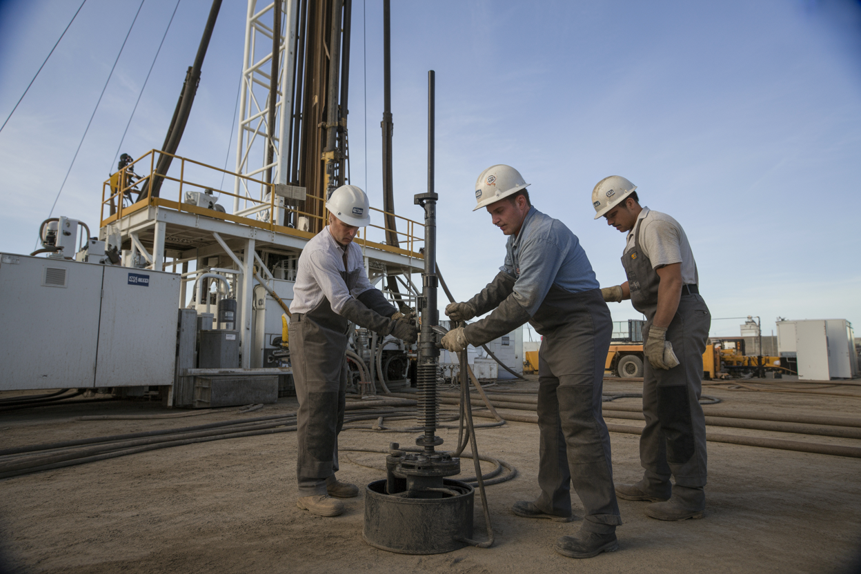 Create a realistic image of skilled technicians installing and setting up drilling fluid management equipment at an onshore drilling rig site, showing white and black male workers in hard hats and safety gear carefully positioning mud pumps, shale shakers, and storage tanks with precision, surrounded by industrial drilling equipment and machinery, with proper lighting illuminating the organized installation process against a clear daytime sky, emphasizing efficiency and professional best practices in the setup procedure, absolutely NO text should be in the scene.