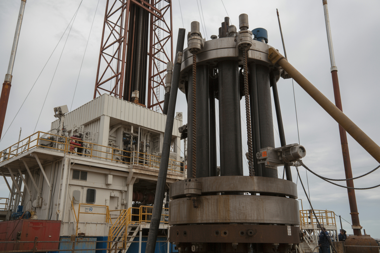 Create a realistic image of a close-up view of a blowout preventer (BOP) stack installed on an onshore drilling rig, showing the large hydraulic rams, control lines, and heavy-duty steel components, with the drilling rig structure visible in the background, set in a rugged oilfield environment under overcast industrial lighting that emphasizes the mechanical complexity and critical safety nature of the equipment, Absolutely NO text should be in the scene.